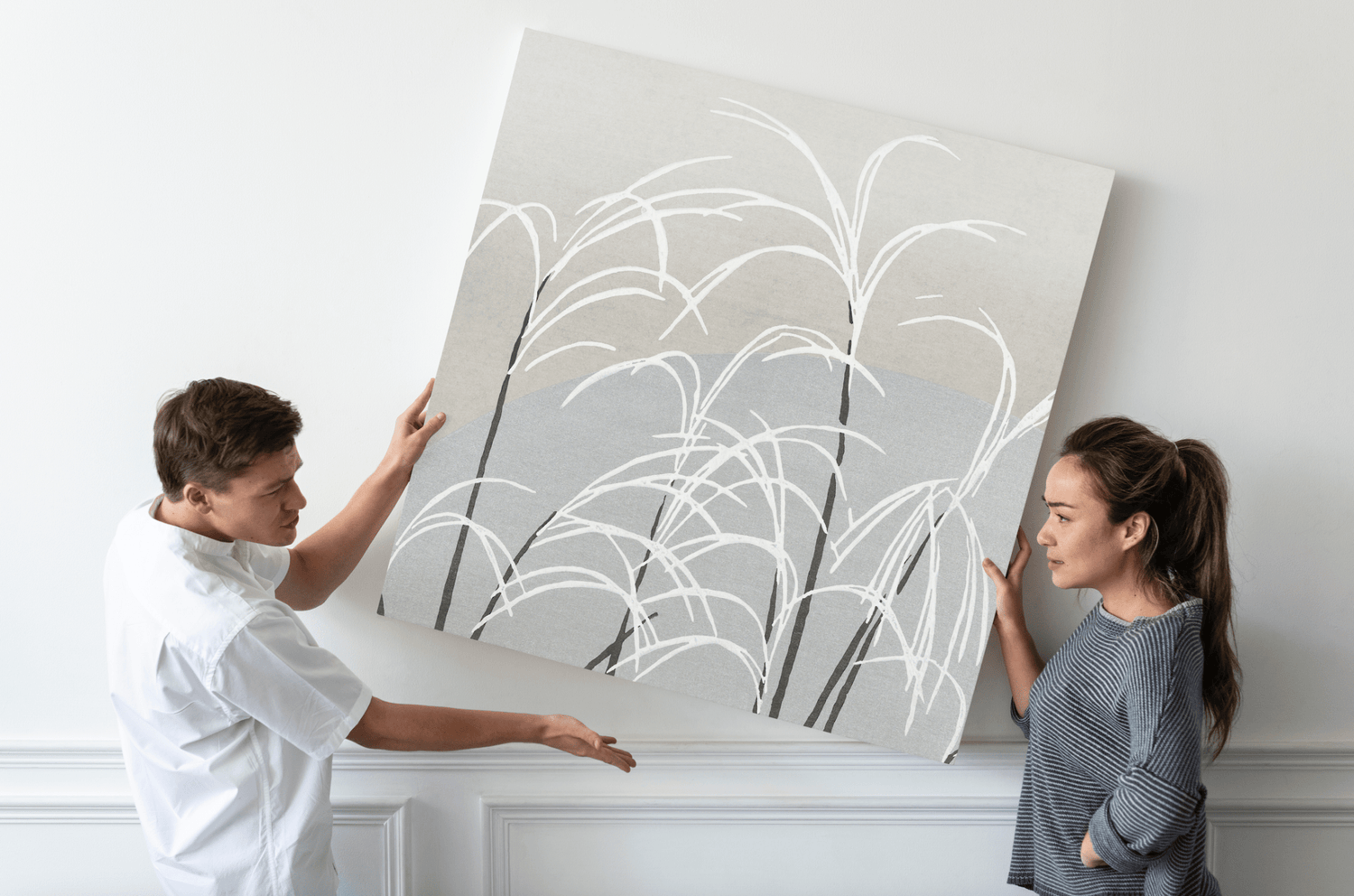 Deux personnes tenant une grande toile d'art abstrait avec des motifs de plantes sur un mur blanc