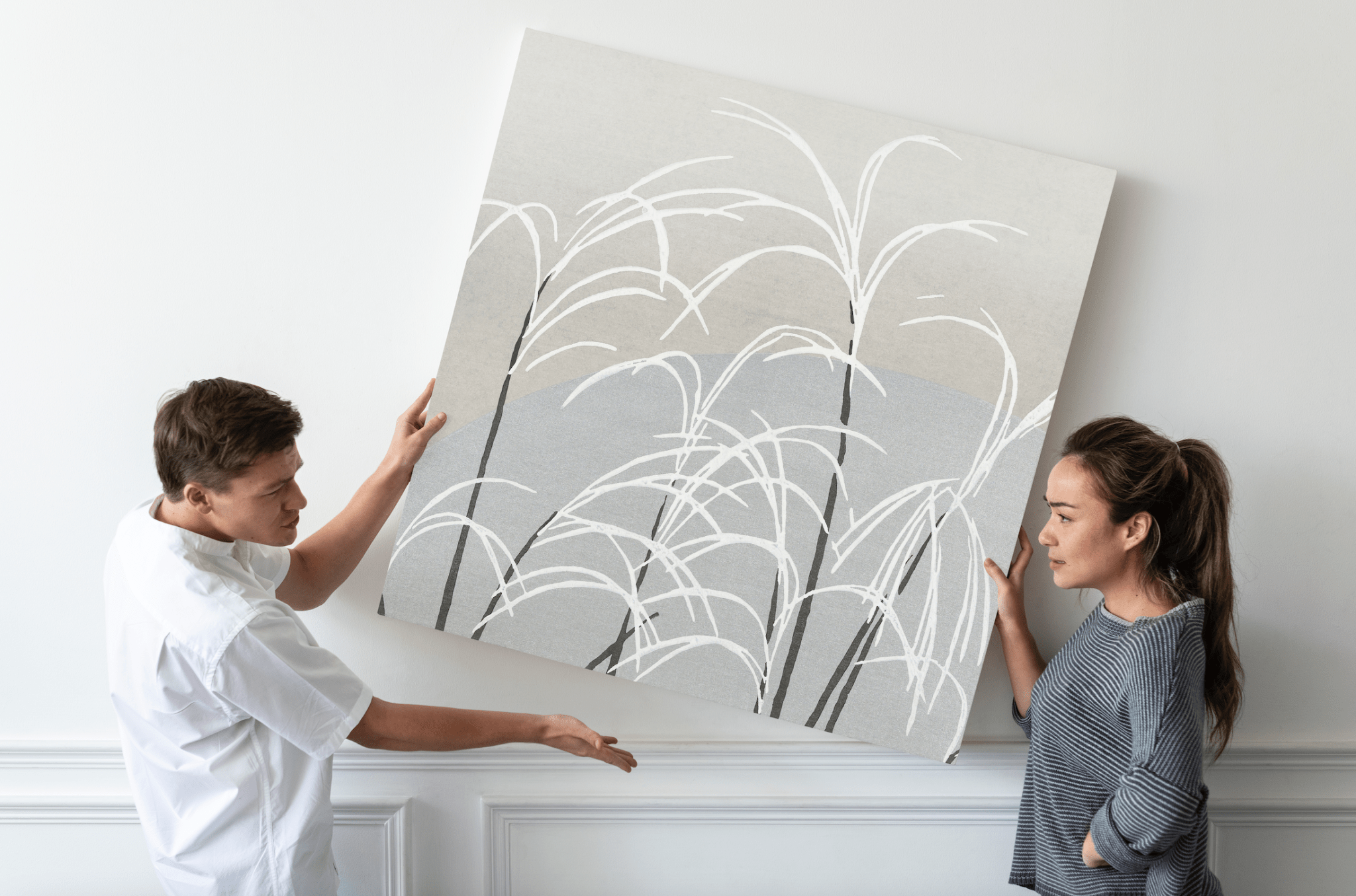 Deux personnes tenant une grande toile d'art abstrait avec des motifs de plantes sur un mur blanc