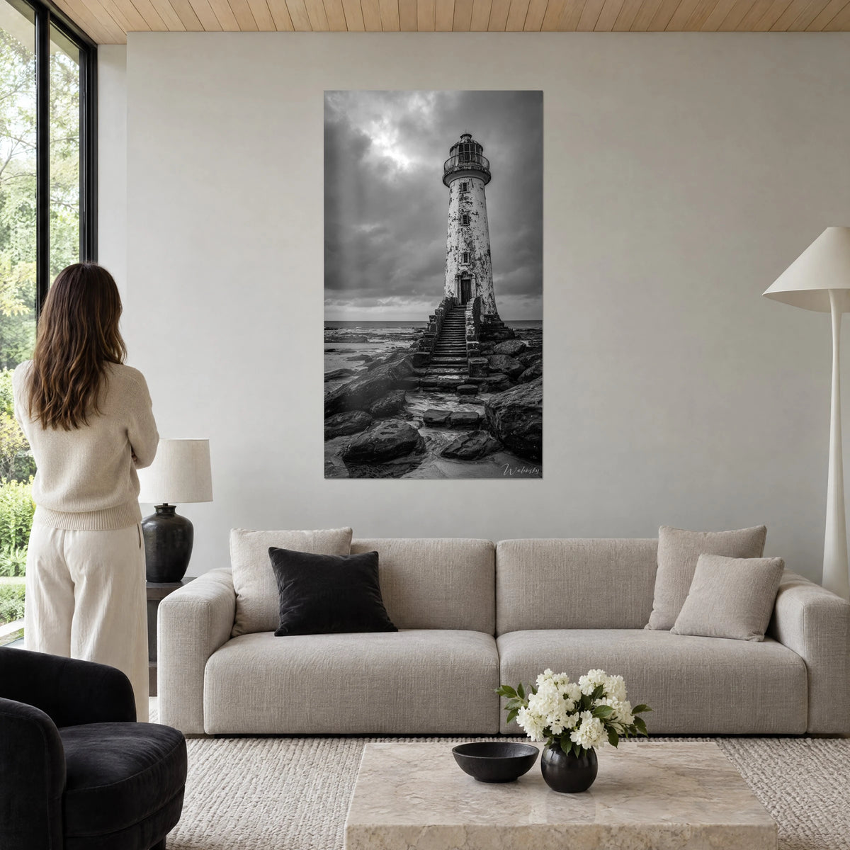 Schilderij oude vuurtoren in zwart-wit, afbladderende toren, stormachtige grijze lucht, vrouw van achteren, boven een bank in een lichte woonkamer