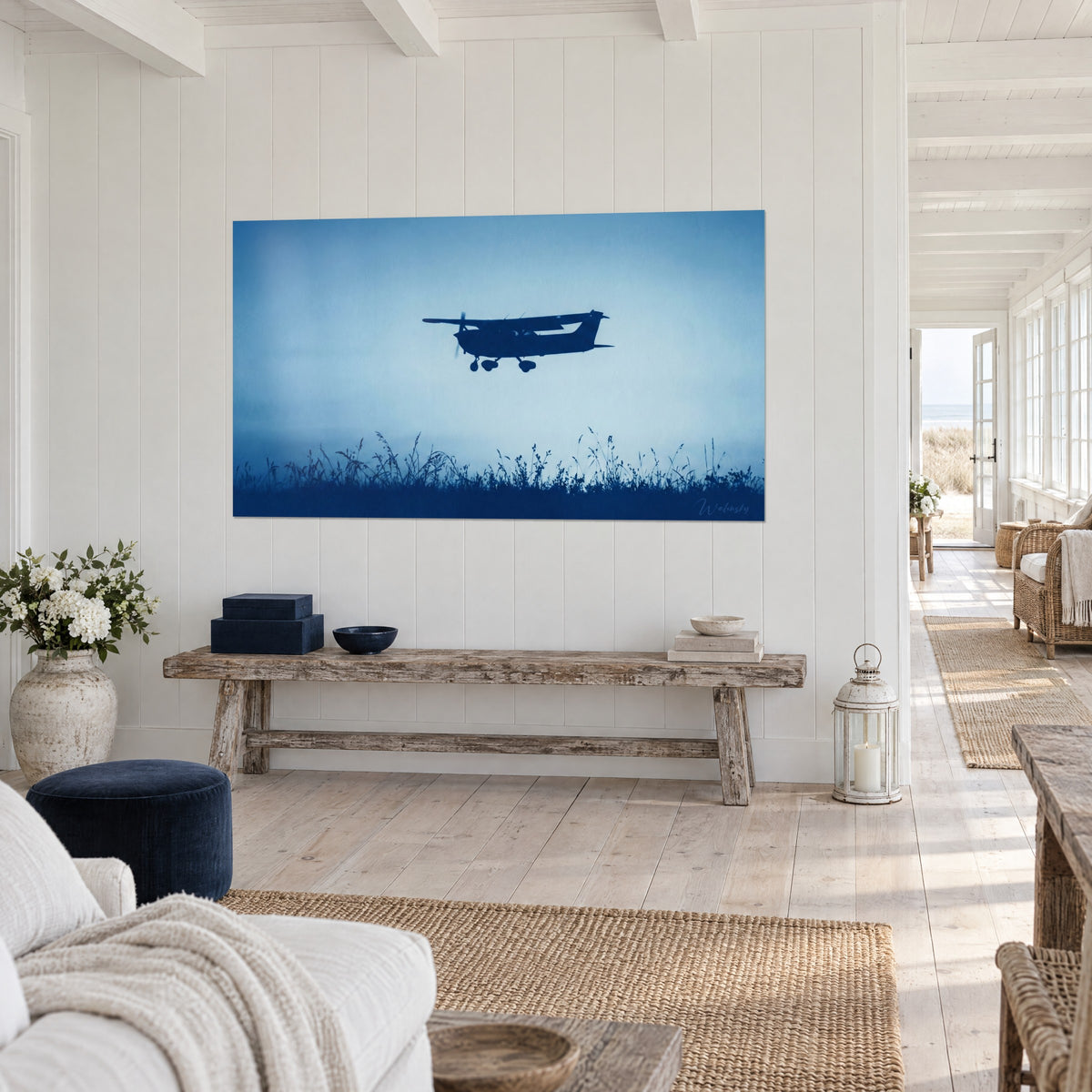 Quadro di un piccolo aereo a elica che sorvola erbe selvatiche, blu notte e azzurro cielo pallido, su una parete bianca perlinata, panca in legno