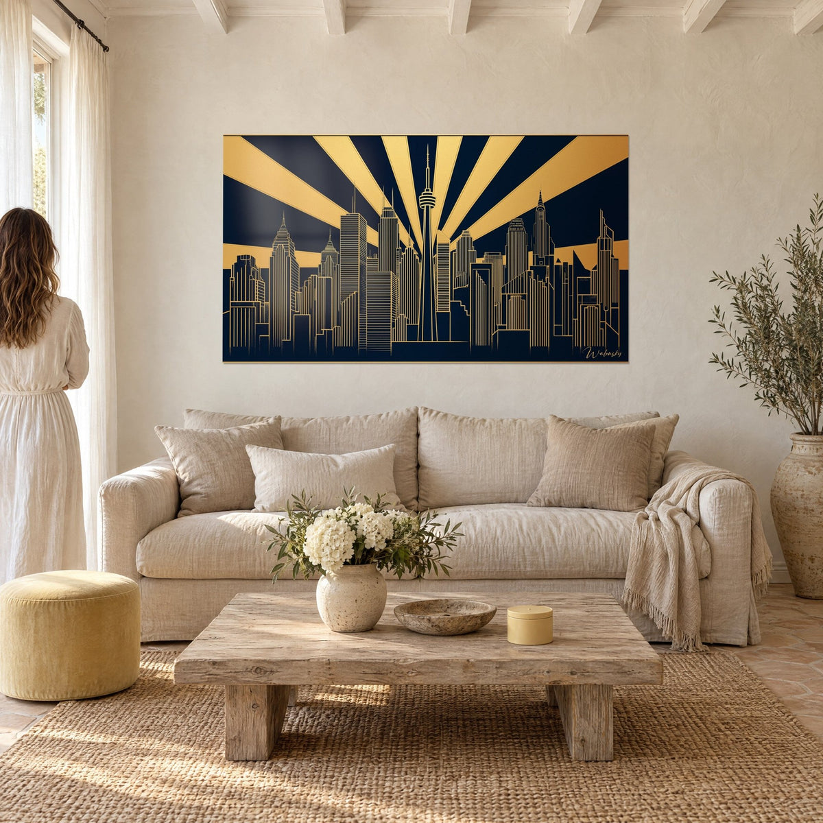 Tableau gratte-ciel stylisés en lignes d'or sur fond bleu nuit, rayons ocre, une femme de dos au-dessus d'un canapé dans un salon lumineux