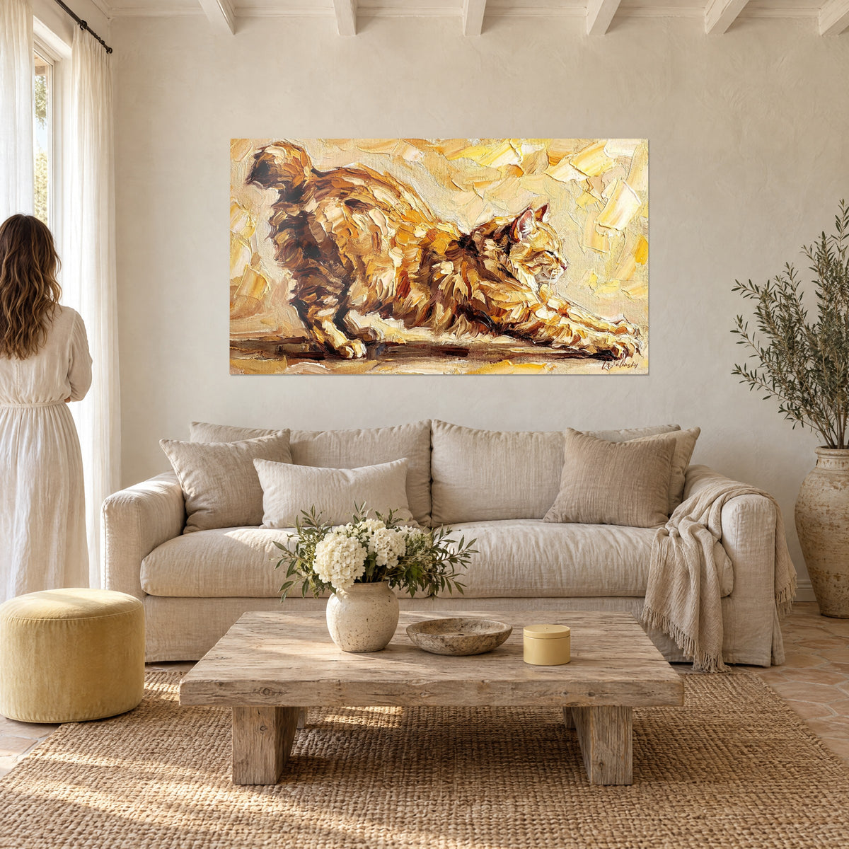 Schilderij van een rossige kat die zich uitrekt in gele en oker spatelstrepen, van achteren bekeken door een vrouw, boven een bank in een lichte woonkamer