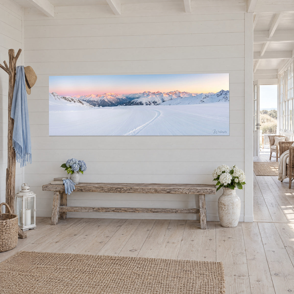 Wall art alpine panorama at dawn, orange-tinged snowy ridges and pale blue sky, on a white wood-paneled wall of a coastal house