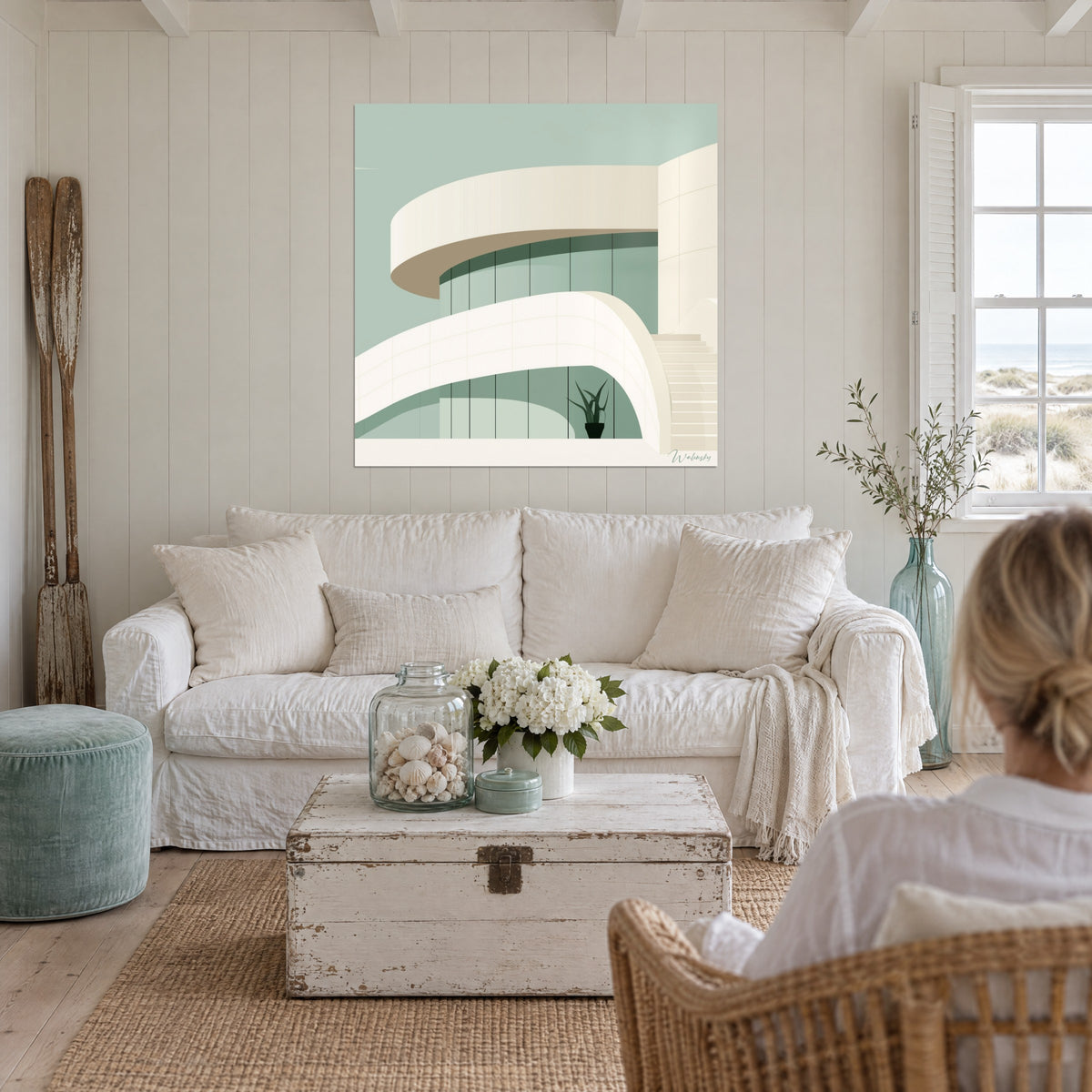 Tableau bâtiment courbe aux terrasses crème sur ciel vert sauge, plante en pot, une personne de dos, au-dessus d'un canapé dans un salon lumineux