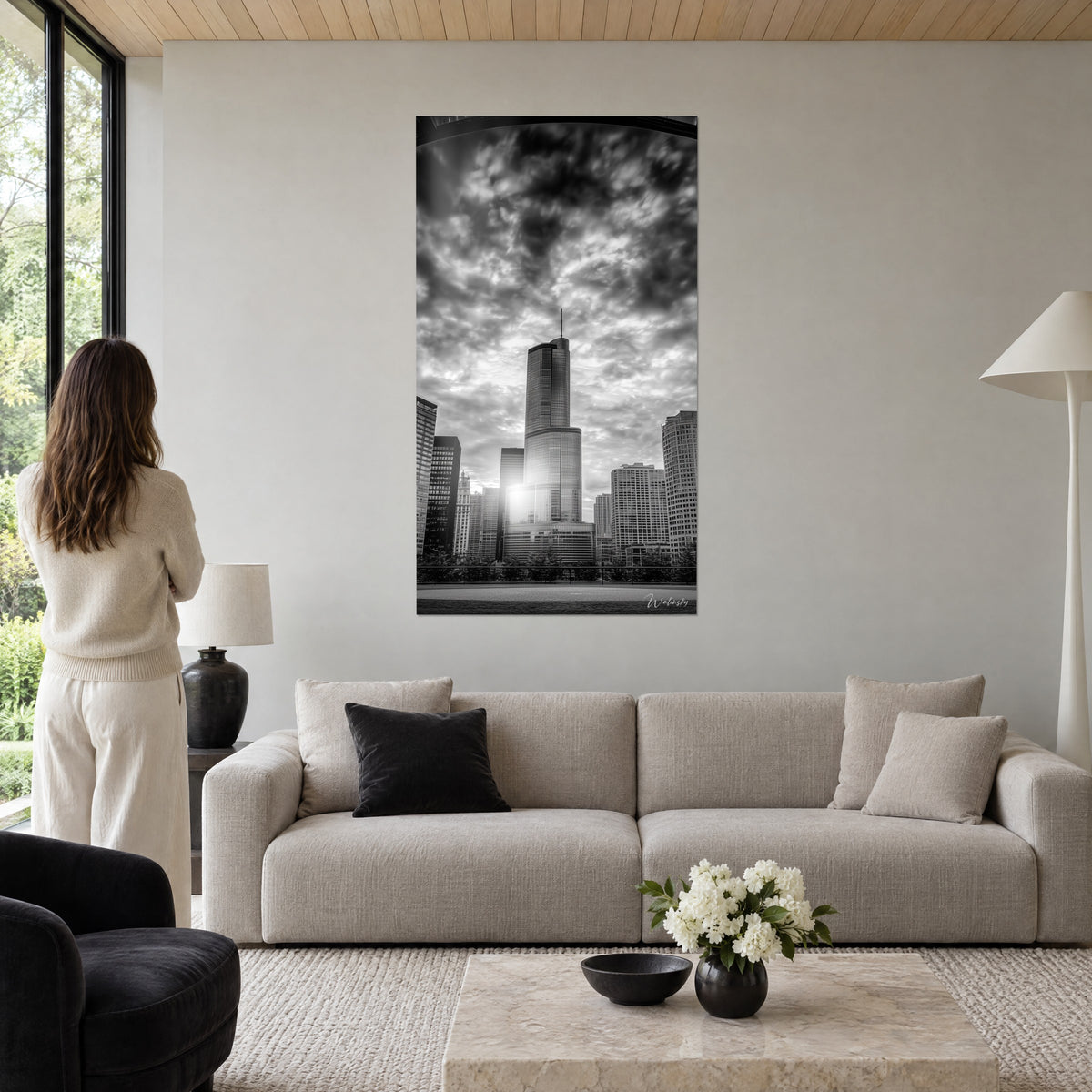 Wall art of a glass skyscraper under turbulent clouds, black and white, white sun at its base, woman seen from behind above a sofa in a bright living room