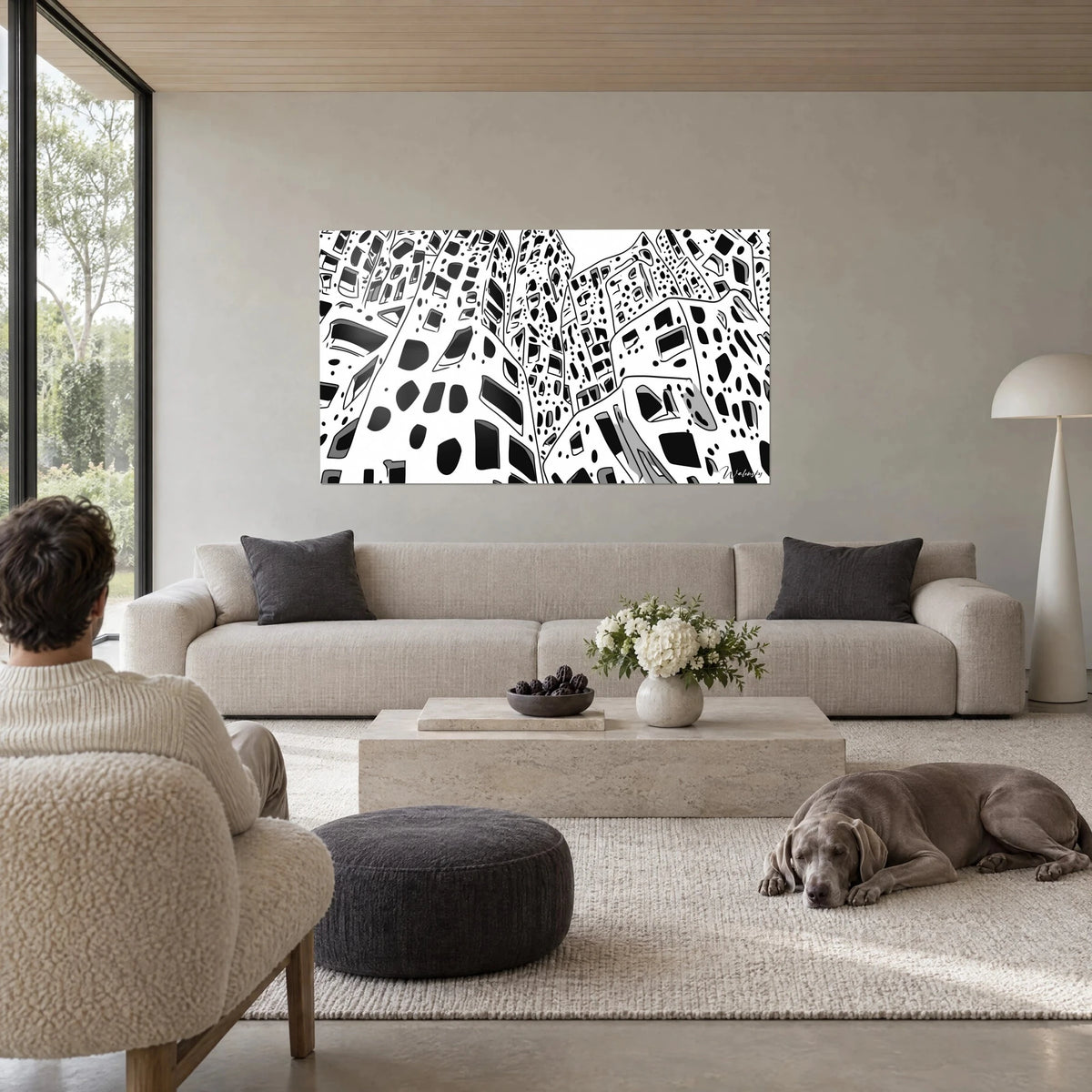 Wall art of perforated skyscrapers with black ovals on a white background, seen from a low angle, two people seen from behind, above a sofa in a bright living room