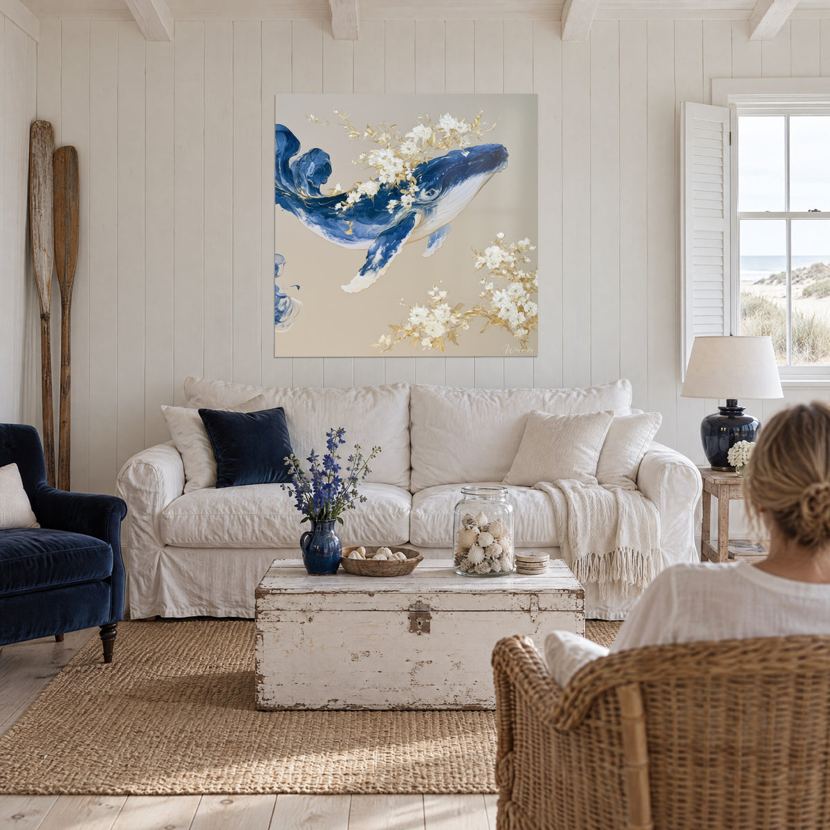 Gemälde eines aufsteigenden Wals in Indigo, umgeben von elfenbeinfarbenen und goldenen Blüten vor beigem Hintergrund, über einem Sofa in einem hellen Wohnzimmer