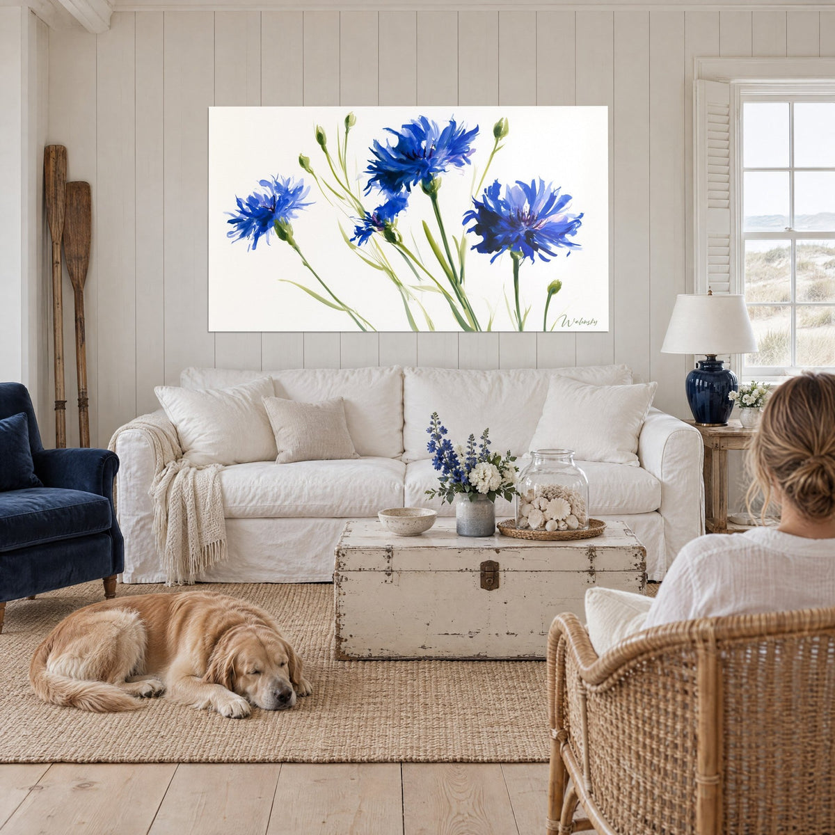 Schilderij korenbloemen in koningsblauw met gefranjerde bloembladen op olijfgroene stengels, witte achtergrond, boven een bank in een lichte woonkamer