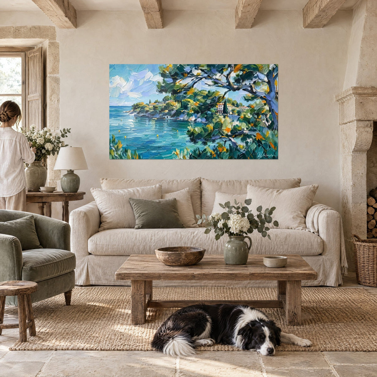 Tableau crique turquoise ourlée de pins, toits ocre et eau bleue, une femme de dos et un collie, au-dessus d'un canapé dans un salon lumineux