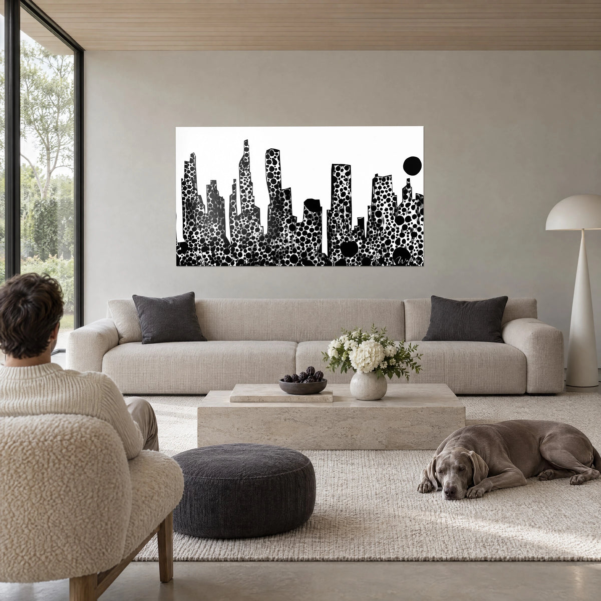 Schilderij wolkenkrabbers met zwarte stippen op witte achtergrond, ronde zon bovenaan, kijker van achteren, boven een bank in een lichte woonkamer
