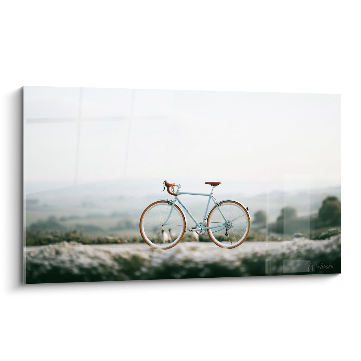 Vélo bleu pastel à selle cognac posé sur route brumeuse, cadre net et fond flou vert-gris