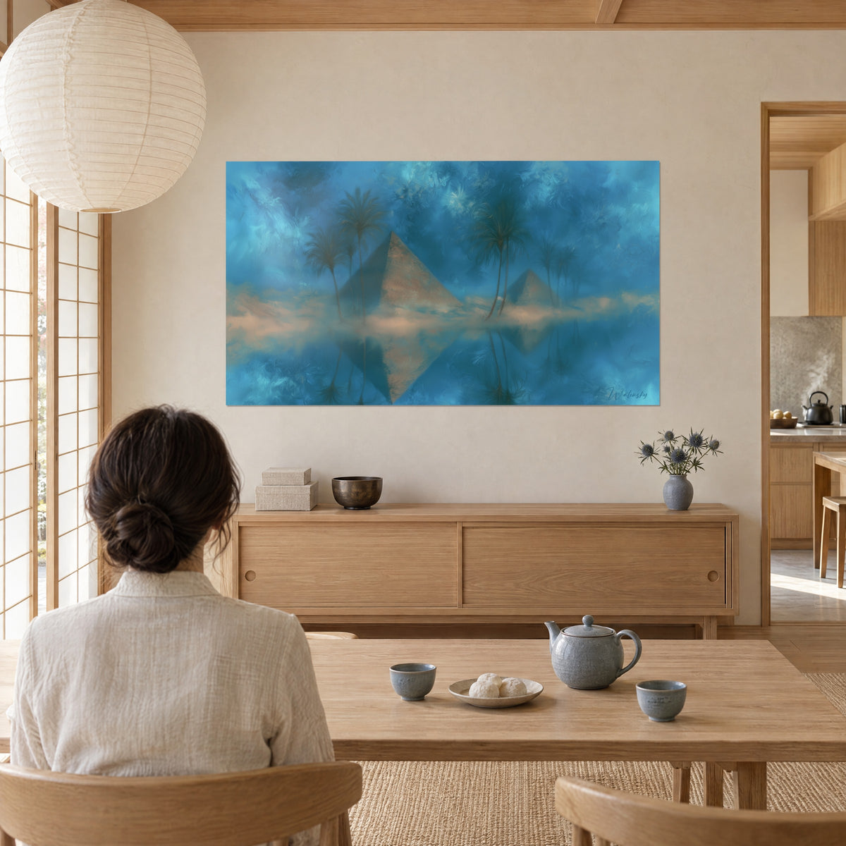 Tableau paysage du Nil onirique, pyramide beige rosé et palmiers bleu-vert, accroché dans un séjour japandi où une femme prend le thé