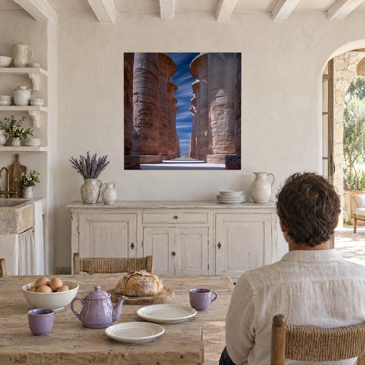 Tableau passage entre piliers de pierre rosée gravés d'inscriptions, ciel indigo, homme de dos attablé dans une cuisine provençale
