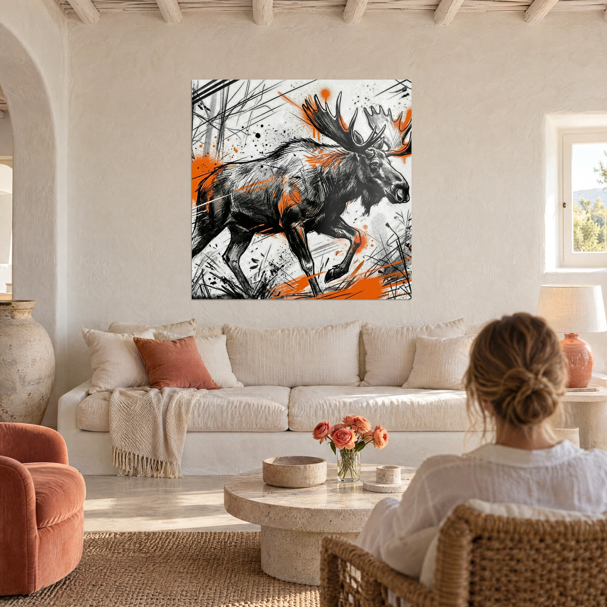 Wall art massive elk with black antlers splashed with bright orange, woman seen from behind, above a sofa in a bright living room