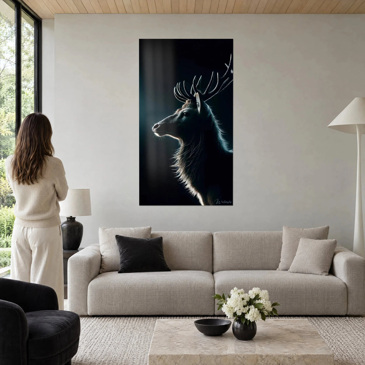 Wall art backlit stag, branched antlers and silver mane on black-green background, woman seen from behind, above a sofa in a bright living room