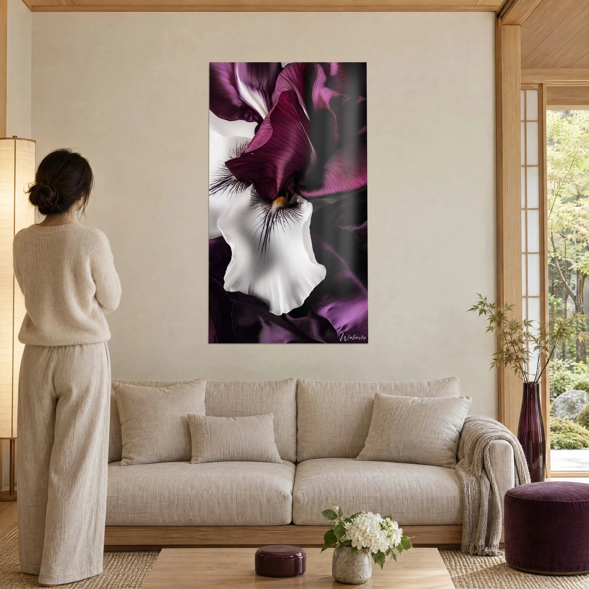 Schilderij witte iris met violette aderen op satijnen pruimkleurige bloembladen, vrouw met de rug naar de kijker, boven een bank in een lichte woonkamer