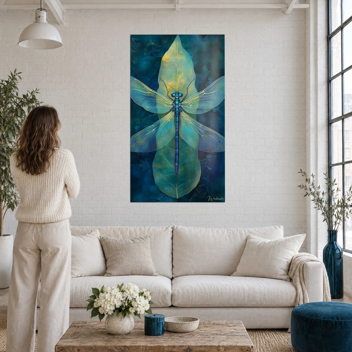 Schilderij libel met kanten vleugels in turquoise en goud op een groen blad, een vrouw van achteren, boven een bank in een lichte woonkamer