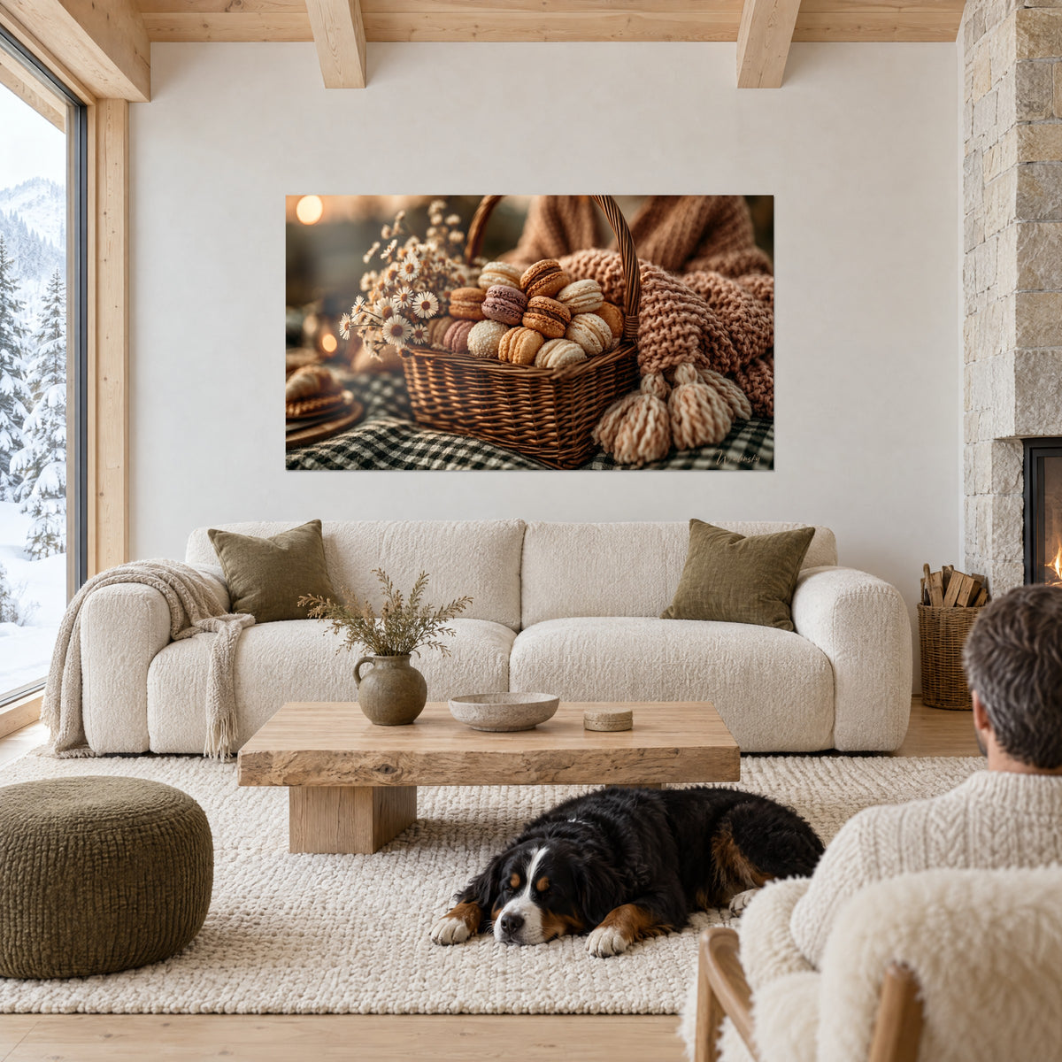 Schilderij mandje van riet gevuld met beige en roze macarons, witte margrieten, gebreide plaid, slapende hond, boven een bank in een lichte woonkamer