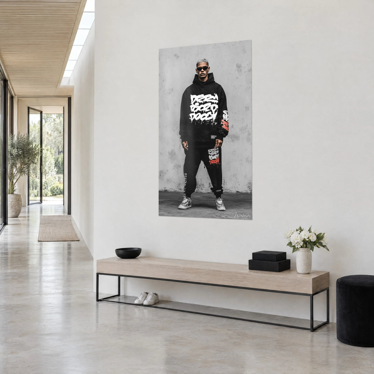 Cuadro retrato streetwear de un hombre de pelo plateado, pantalón de chándal negro salpicado de blanco y rojo, pared gris, pasillo luminoso en tres cuartos