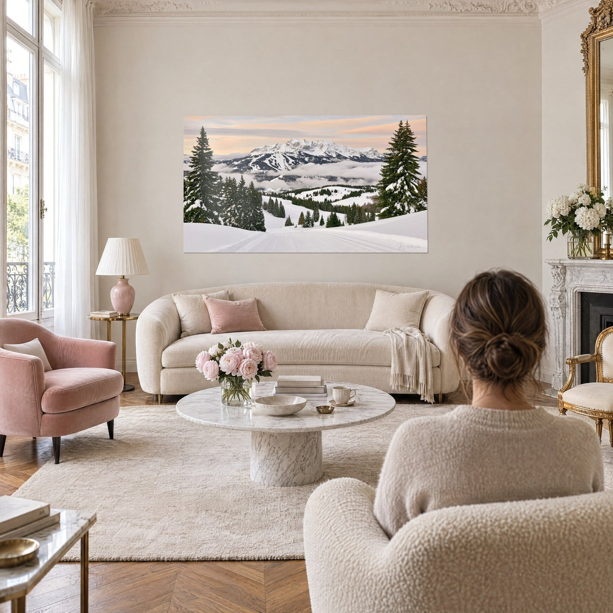 Tableau piste de ski damée bordée de sapins enneigés, ciel rose pêche, femme de dos, au-dessus d'un canapé dans un salon lumineux