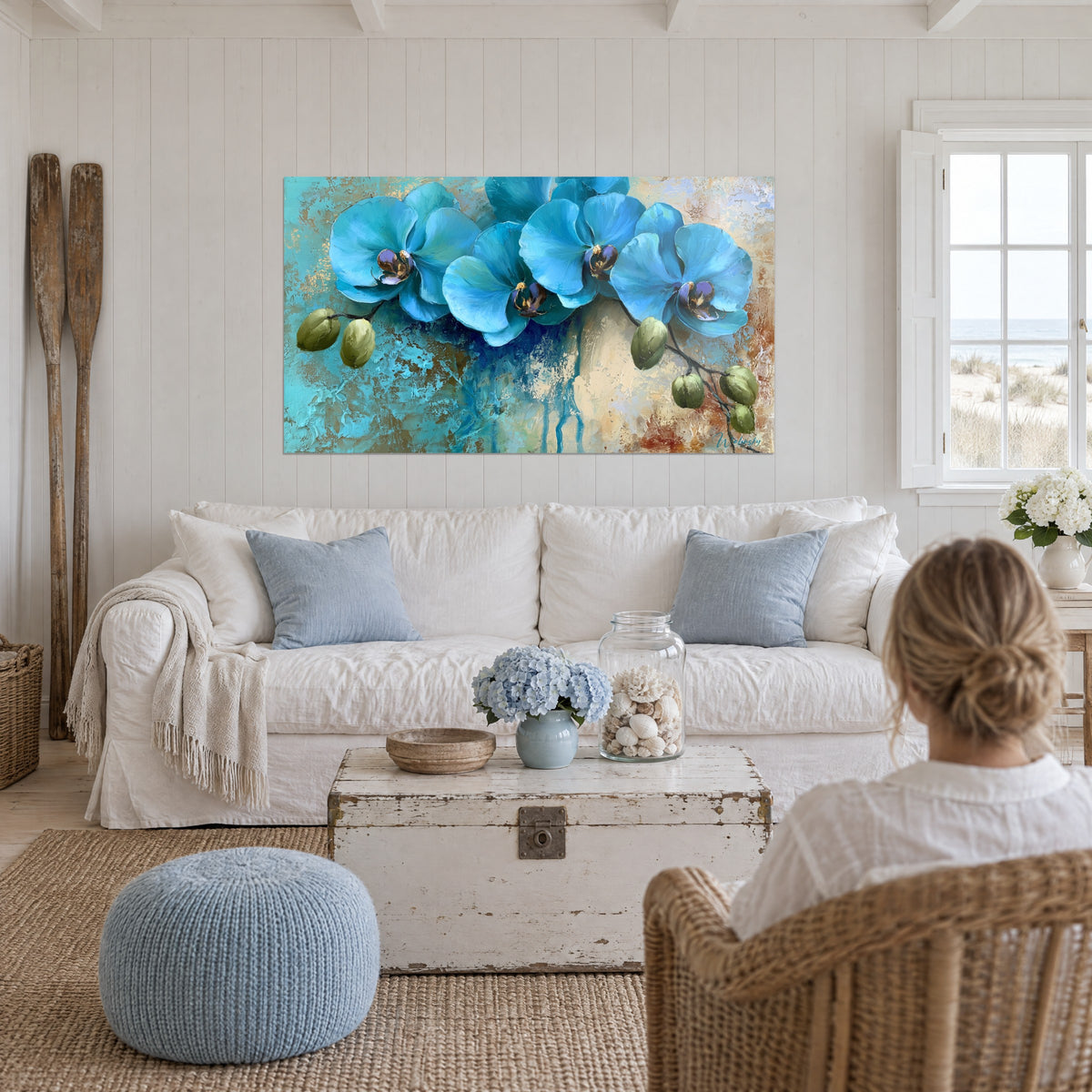 Schilderij orchideeën turquoiseblauw met paarse harten op getextureerde okergele achtergrond, vrouw met de rug naar de kijker boven een bank in een lichte woonkamer