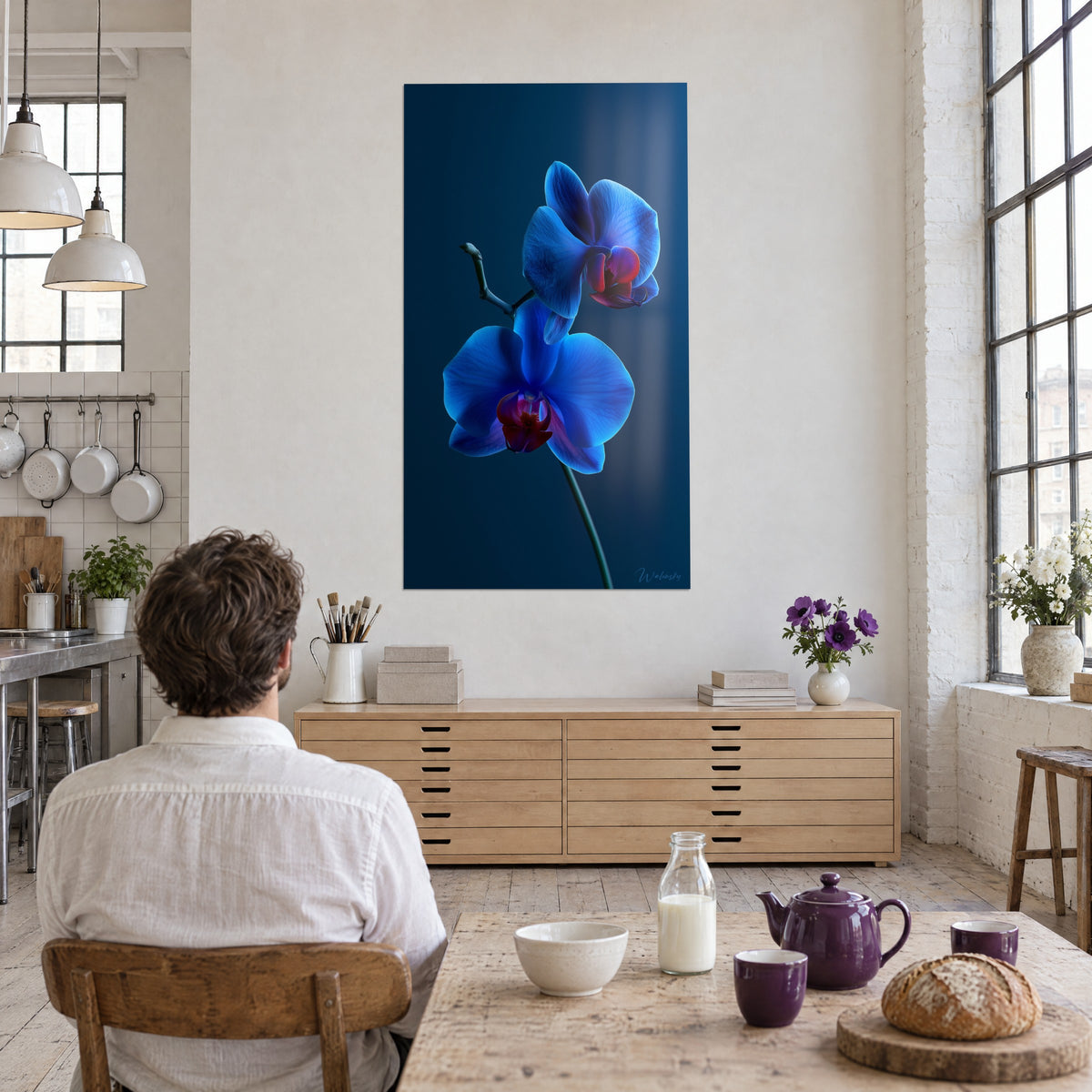 Cuadro orquídea azul cobalto y granate sobre fondo azul marino, hombre de espaldas sentado ante una mesa de desayuno, loft de paredes de ladrillo blanco