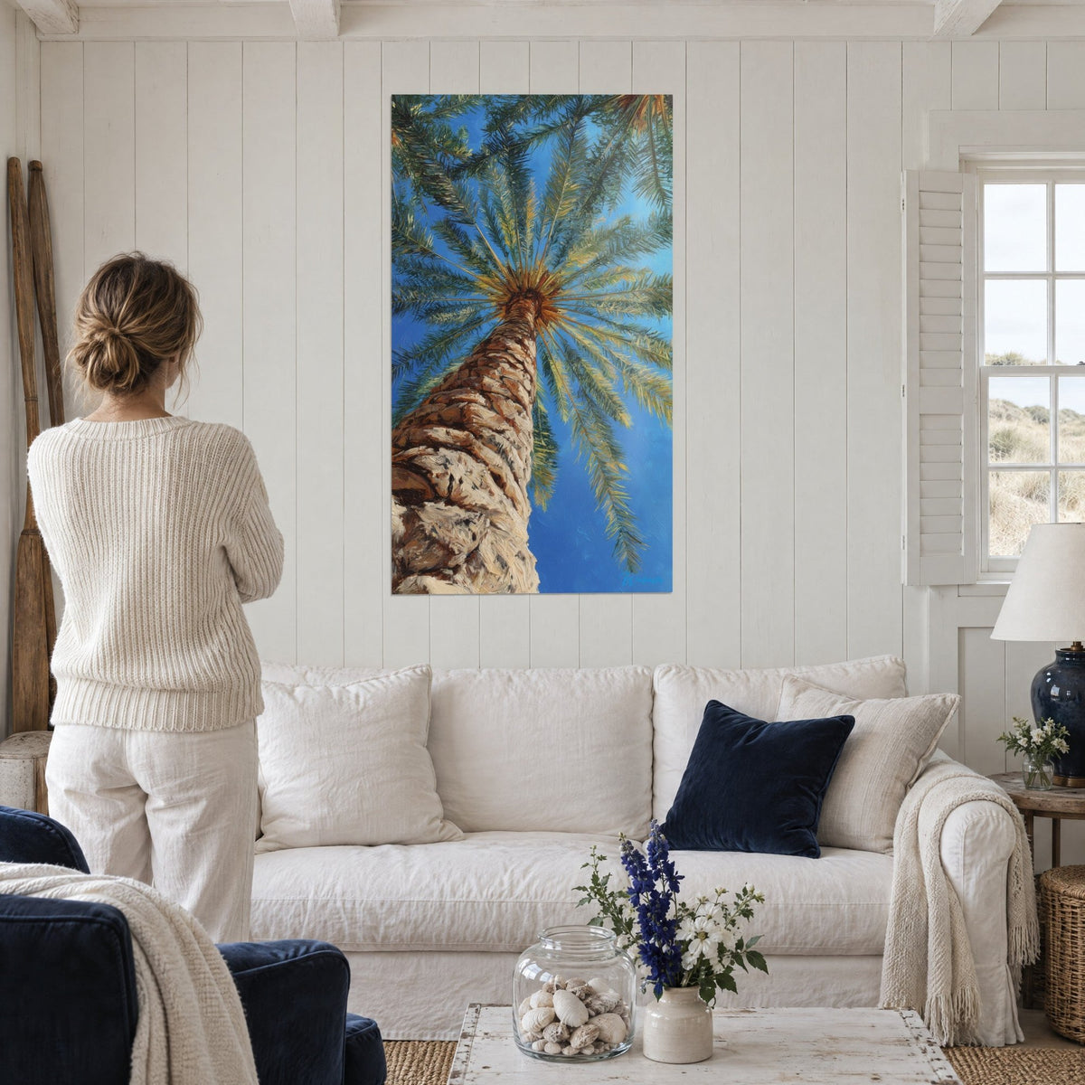 Gemälde Dattelpalme von unten gesehen, brauner Stamm und grüne Palmwedel vor blauem Himmel, über einem Sofa in einem hellen Wohnzimmer