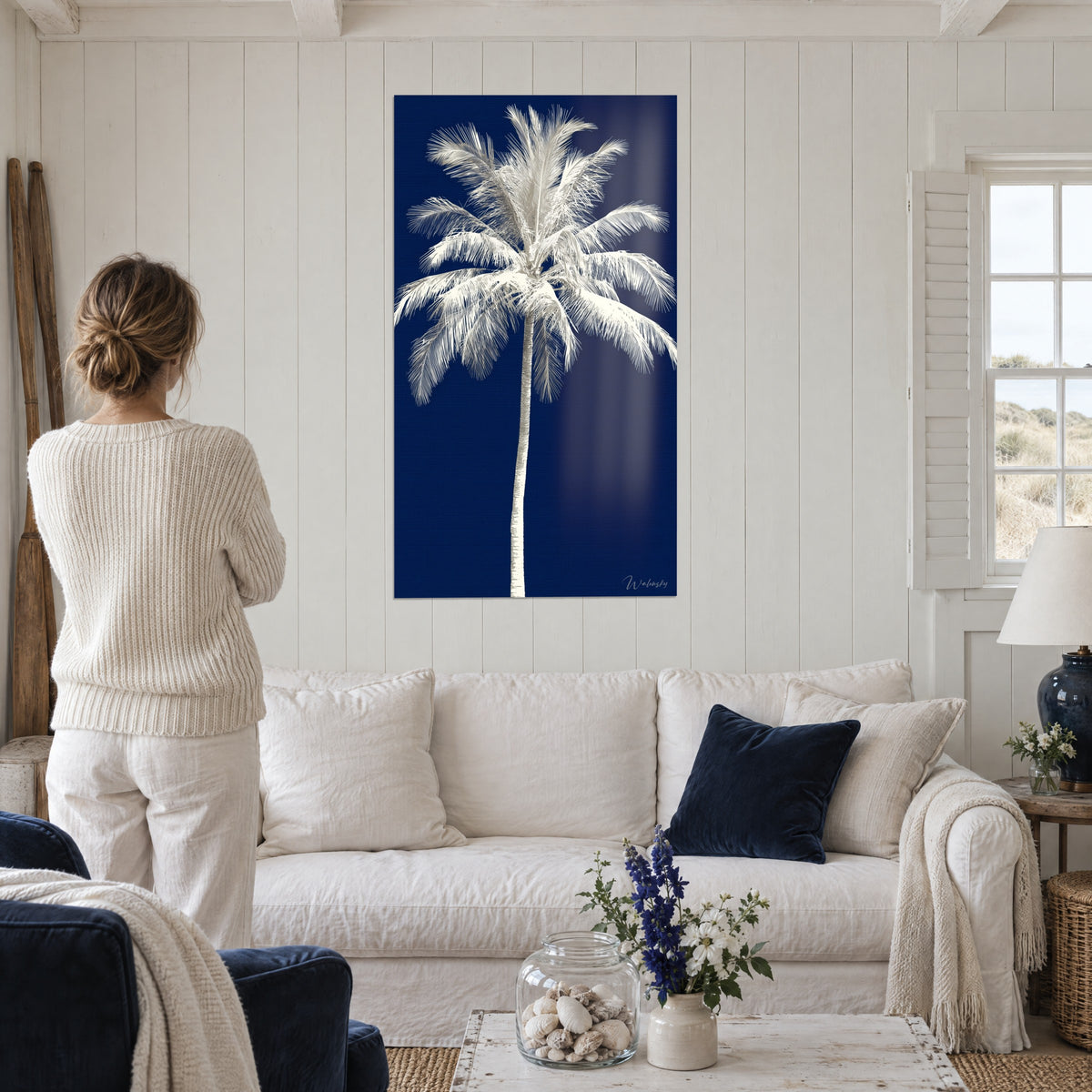 Tableau palmier ivoire aux palmes retombantes sur fond bleu marine, femme de dos, au-dessus d'un canapé dans un salon lumineux