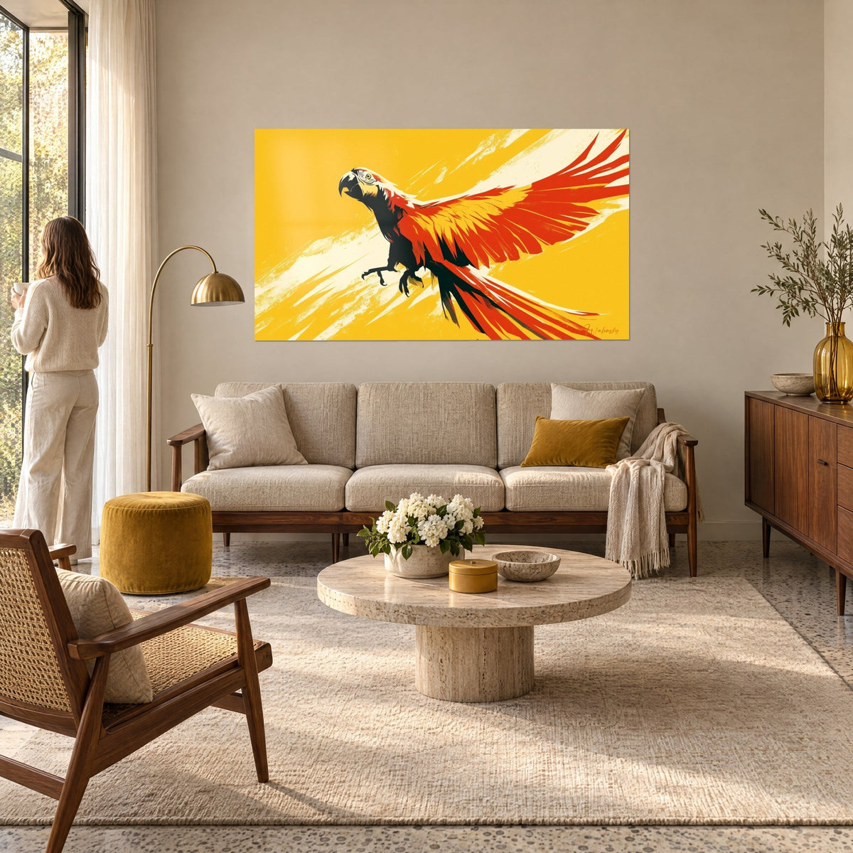 Schilderij scharlaken ara met uitgespreide vleugels, rode en gouden veren op een zonnig geel achtergrond, een vrouw van achteren boven een bank in een lichte woonkamer