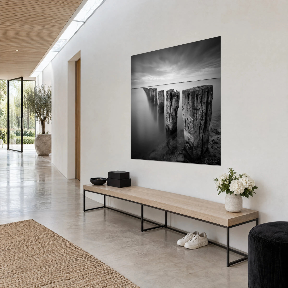 Tableau alignement de poteaux marins en noir et blanc, bois fissuré gris cendré, ciel strié de nuages, en vue de trois-quarts dans un couloir clair
