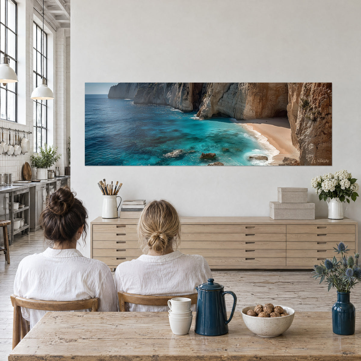 Wall art of a wild shoreline where white foam fringes turquoise water beneath a golden-brown rock, two people seen from behind beside a table
