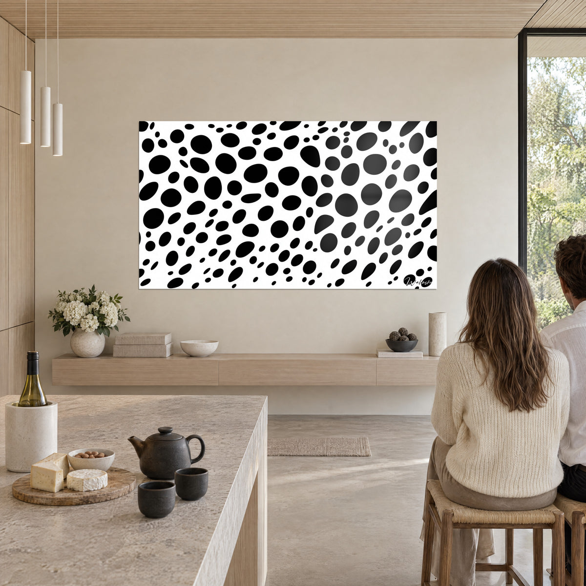 Tableau constellation de pastilles noires irrégulières sur blanc, couple de dos près d'un îlot en pierre beige, cuisine ouverte et théière noire
