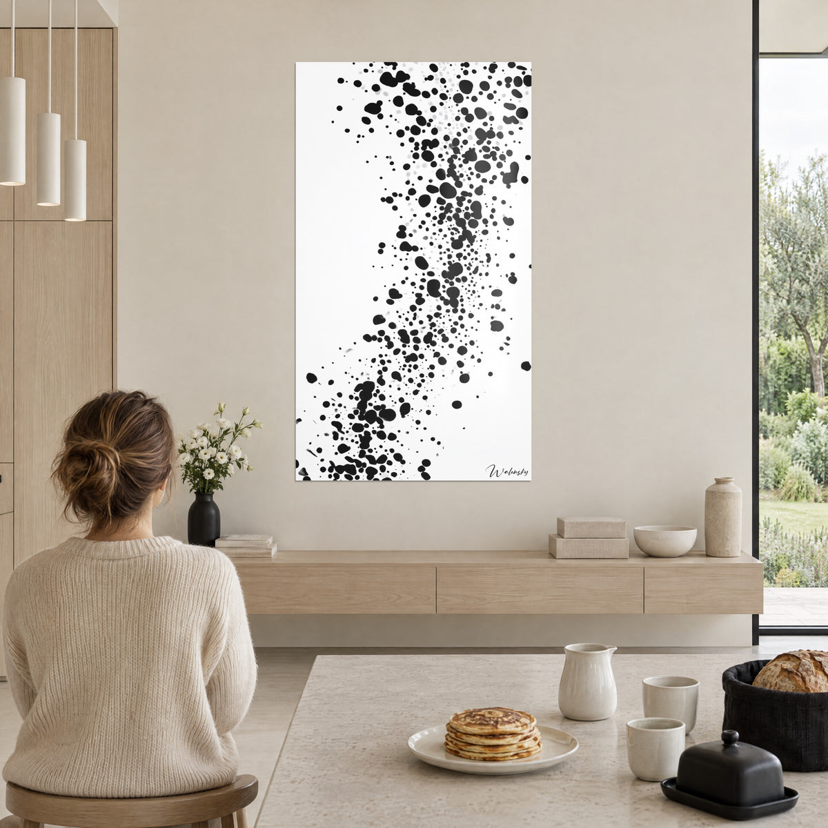 Cuadro con lluvia de gotas negras y grises sobre blanco, mujer con moño suelto de espaldas en la mesa, tortitas y pared crema
