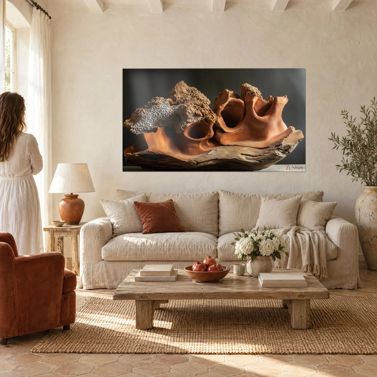 Tableau sculpture d'argile orange et corail grisâtre sur bois flotté, vue par une femme de dos, au-dessus d'un canapé dans un salon lumineux