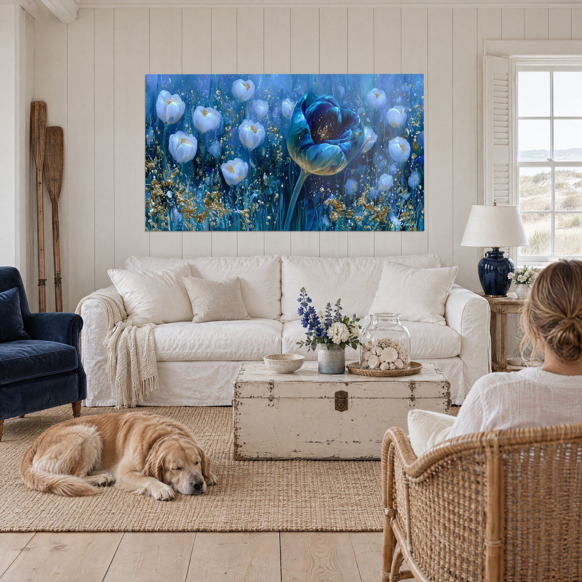 Schilderij van een reusachtige blauwe tulp omringd door witte bloemen met gouden glans, een vrouw met haar retriever, boven een bank in een lichte woonkamer
