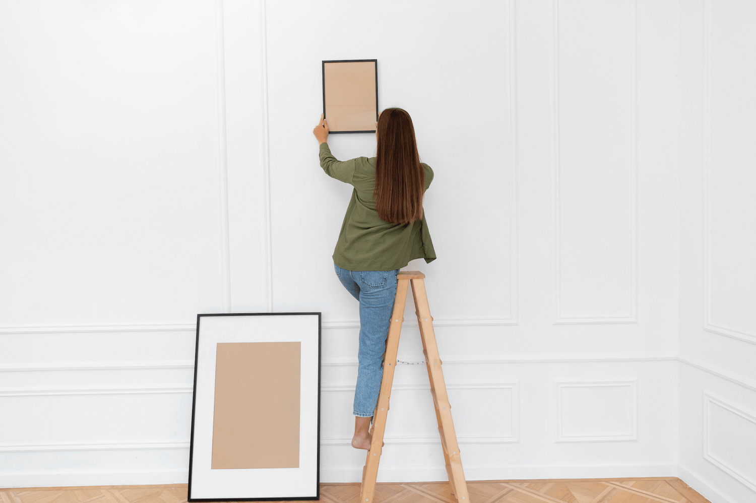 Femme accrochant un cadre vide sur un mur blanc avec un escabeau en bois