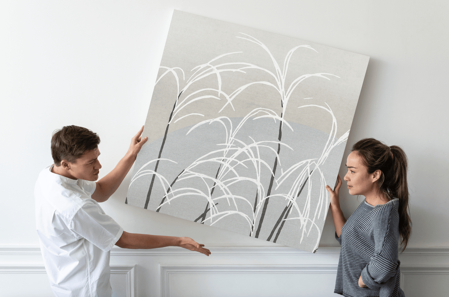 Deux personnes tenant une grande toile d'art abstrait avec des motifs de plantes sur un mur blanc