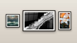 Pared de galería con tres cuadros enmarcados: Playa vista aérea, Tormenta oceánica y Lirio N°2