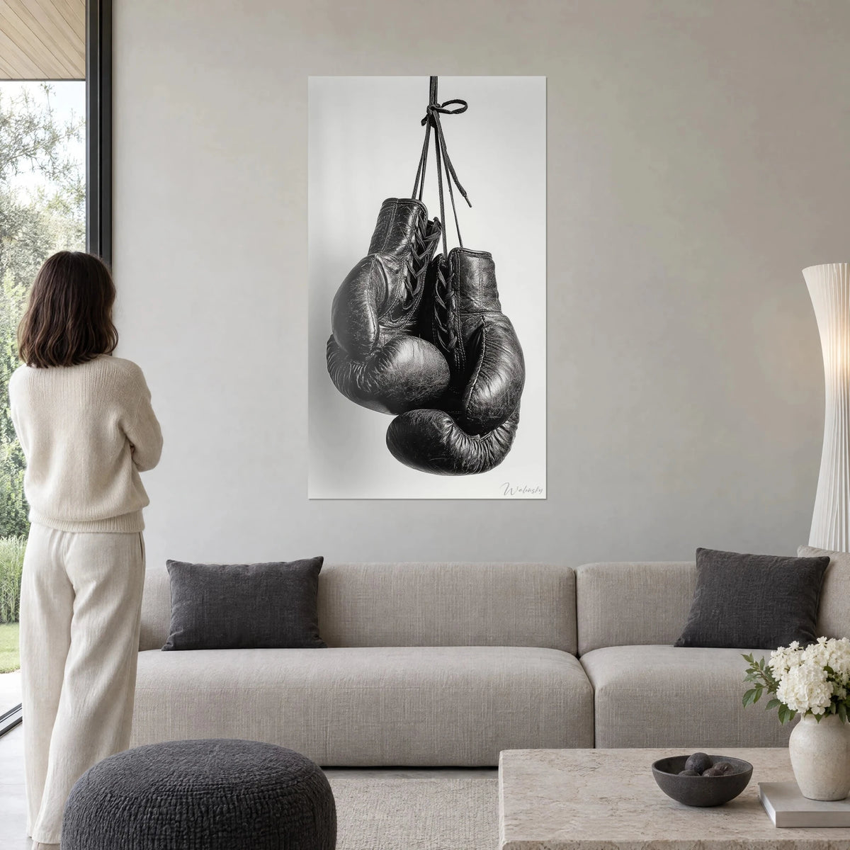 Tableau gants de boxe en cuir noir craquelé, lacets suspendus sur fond gris perle, au-dessus d'un canapé dans un salon lumineux