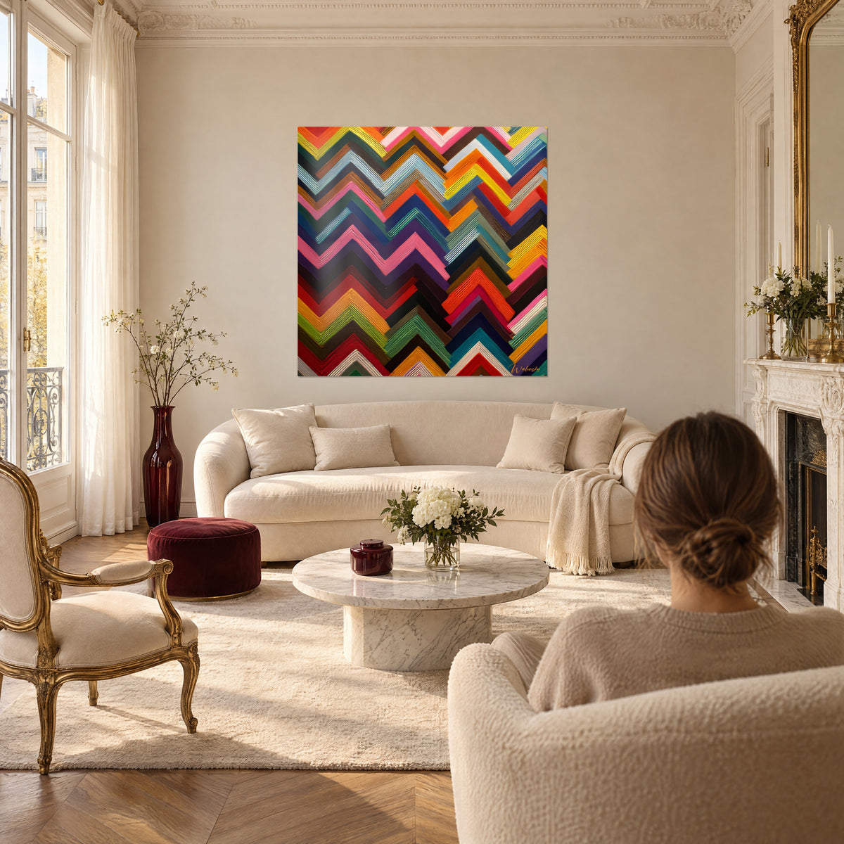 Wall art stacked chevron zigzags in pink, orange and indigo, viewed from behind by a woman above a sofa in a bright living room