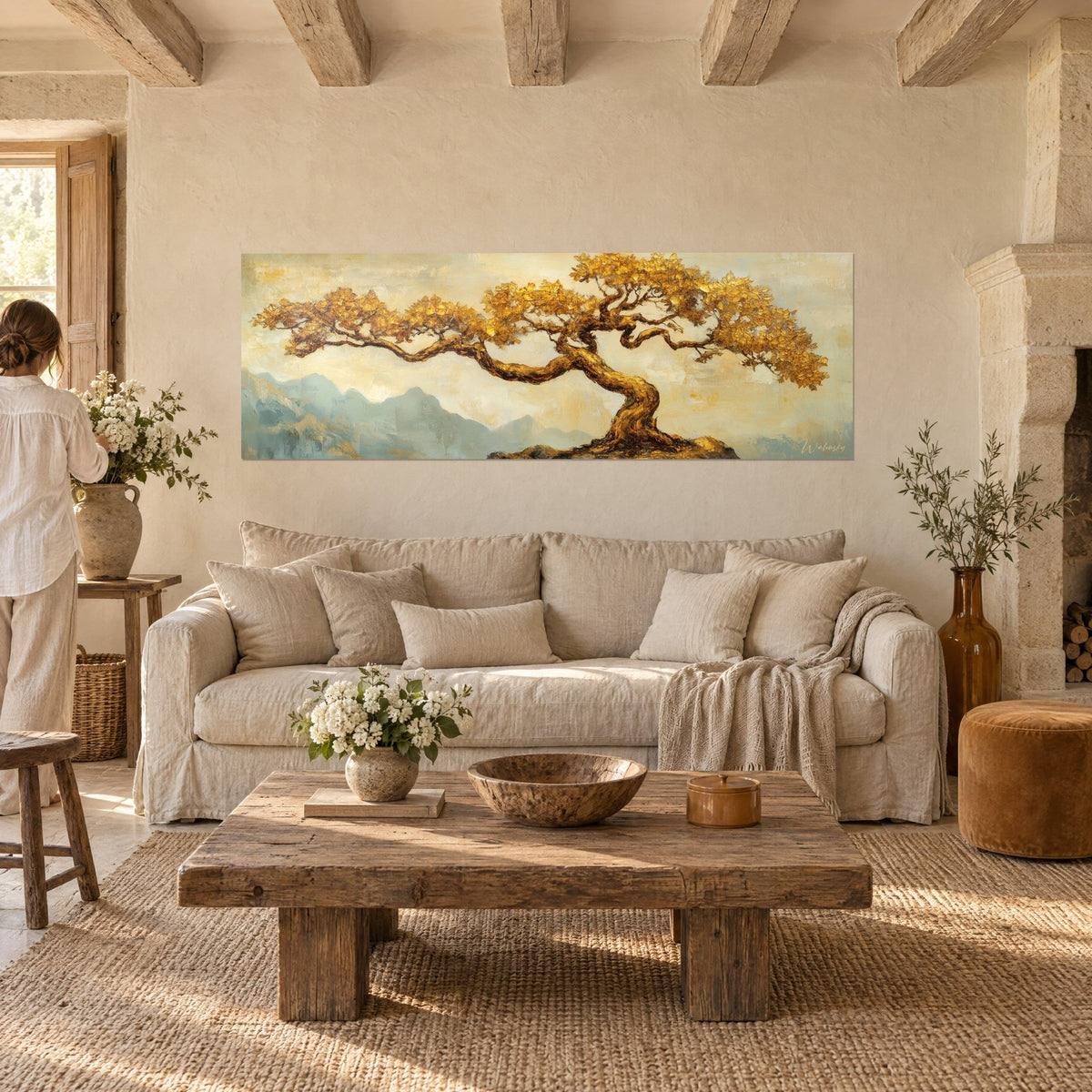 Wall art tree of life with gold leaves in front of blue-grey mountains, woman seen from behind, above a sofa in a bright living room
