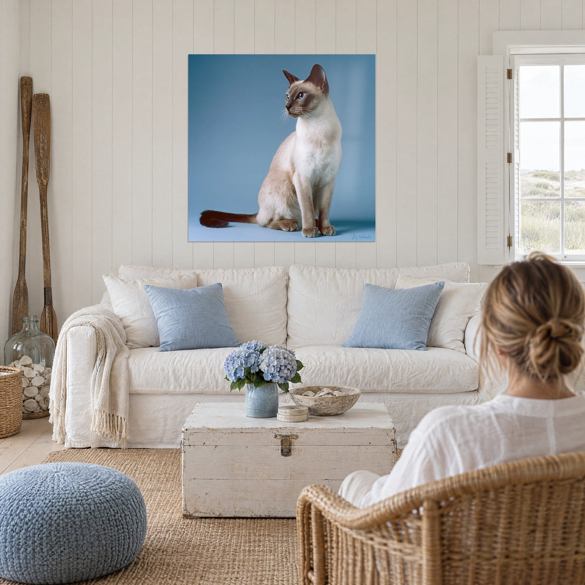 Quadro di un gatto tonkinese seduto, pelo color crema e muso marrone, sfondo blu acciaio, sopra un divano in un soggiorno luminoso