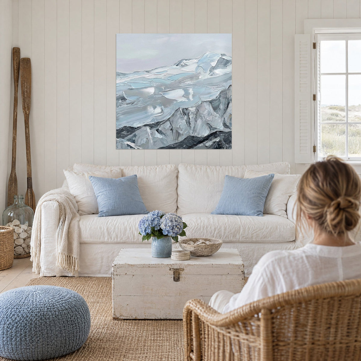Wall art of snowy peaks in thick glacier-blue and steel-grey brushstrokes, a woman seen from behind, above a sofa in a bright living room