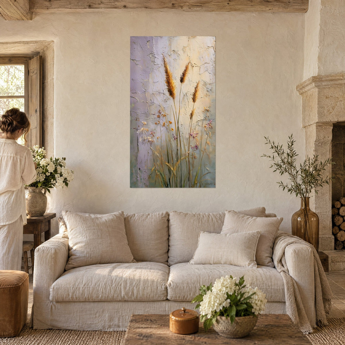 Quadro spighe dorate e fiori rosa pallido su muro screpolato malva e beige, donna di spalle, sopra un divano in un soggiorno luminoso