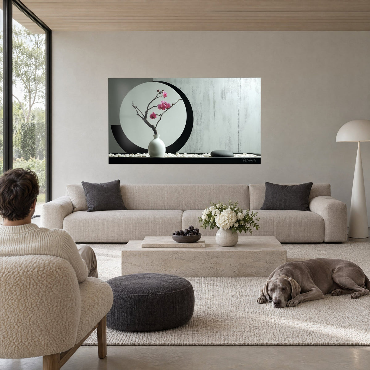 Wall art pink orchid on gnarled branch, white vase and grey disc outlined in black, above a sofa in a bright living room