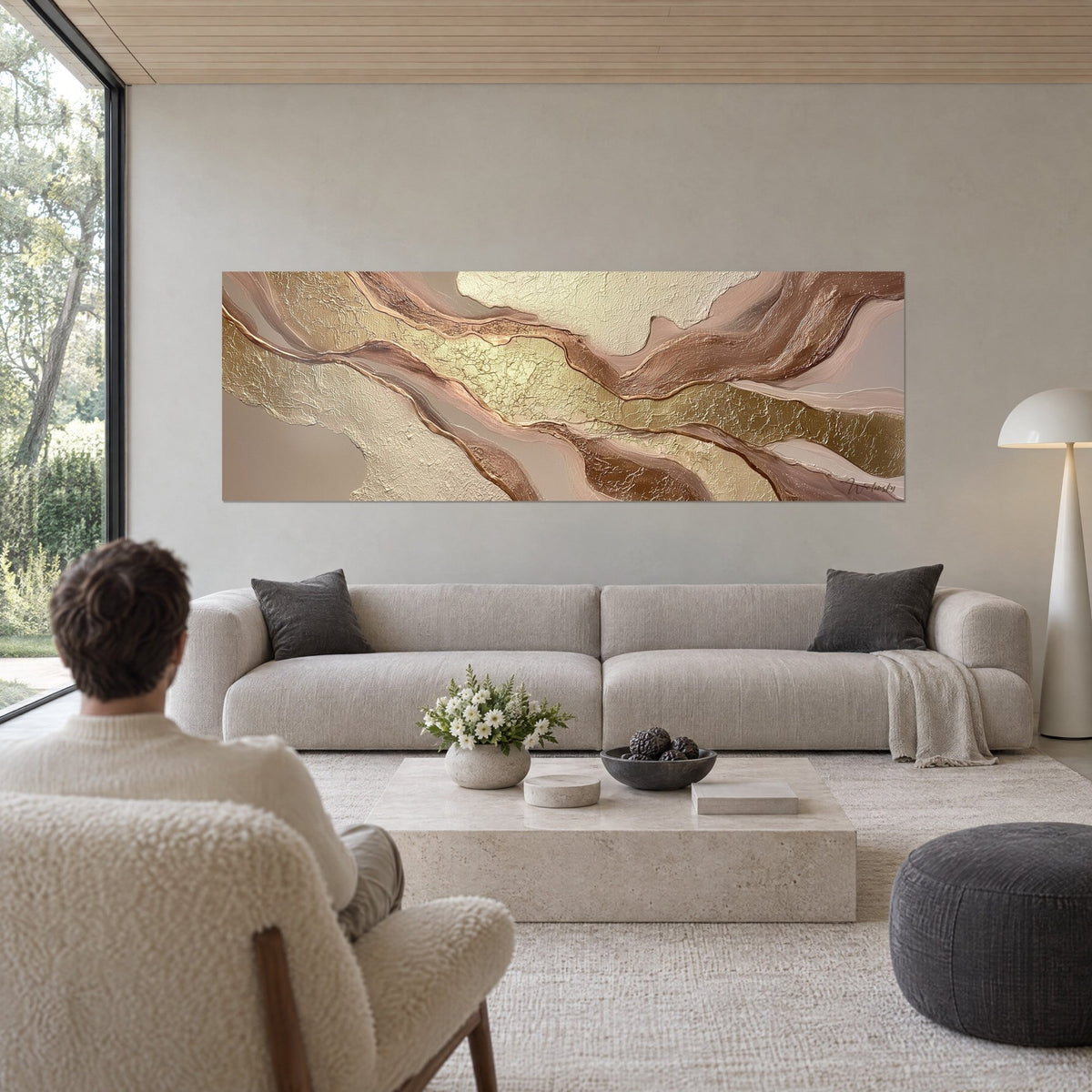 Quadro con nastri di metallo dorato e rame rosato su sfondo beige, visto di spalle da un uomo, sopra un divano in un soggiorno luminoso