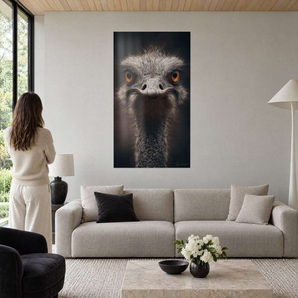 Gemälde Strauß mit durchdringendem orangefarbenem Blick, flauschiges graues Gefieder auf dunkelbraunem Hintergrund, Frau von hinten über einem Sofa in einem hellen Wohnzimmer