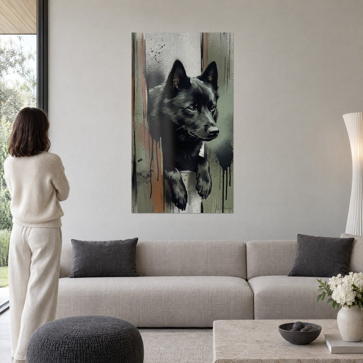 Wall art black Schipperke puppy with amber eyes, sage green background with rust-colored drips, above a sofa in a bright living room