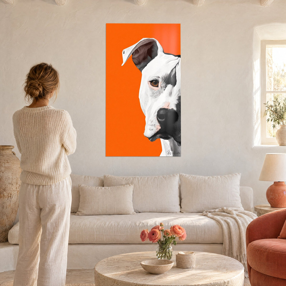 Schilderij witte Dogo Argentino met amberkleurig oog op fel oranje achtergrond, een vrouw in crèmekleurige trui kijkt ernaar boven een bank in een lichte woonkamer