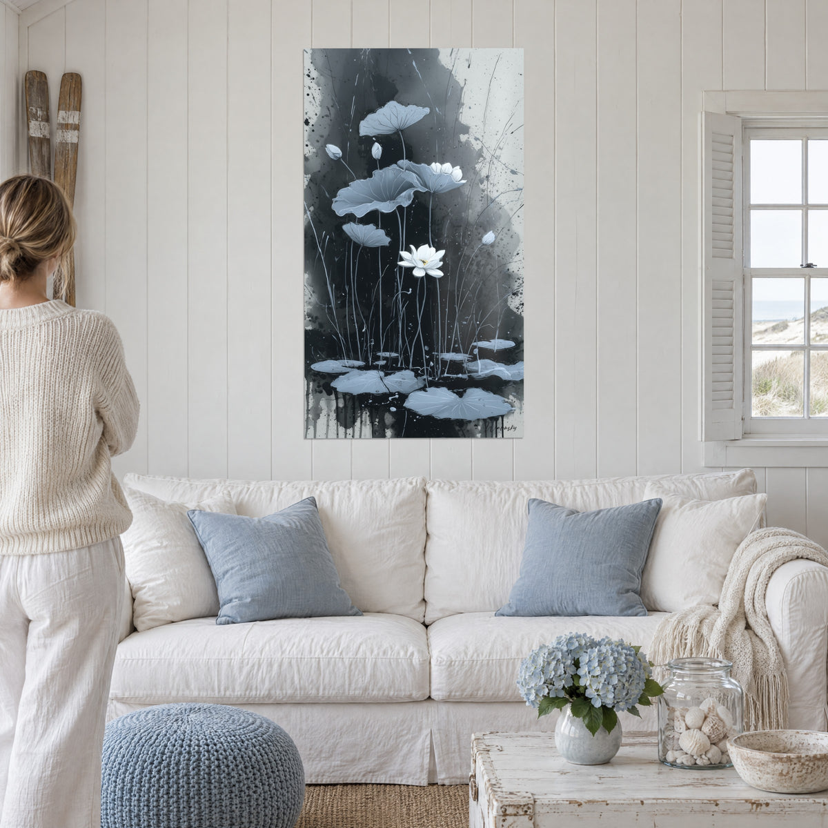 Quadro fiore di loto bianco che si schiude tra foglie grigio-azzurre su fondo di inchiostro nero profondo, sopra un divano in un soggiorno luminoso